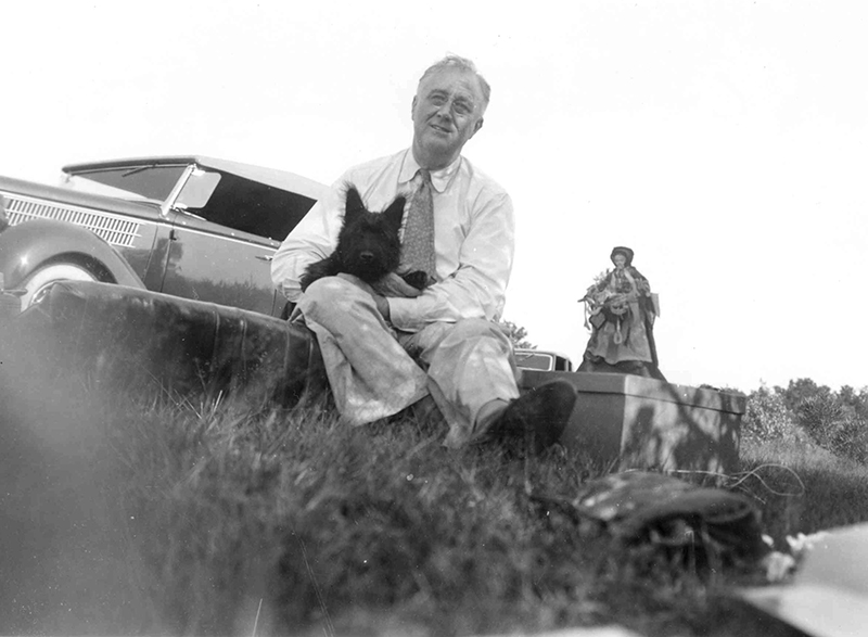 Black-and-white image of man sitting on something in a grassy field, holding a Scottie dog, with a car and another person behind him