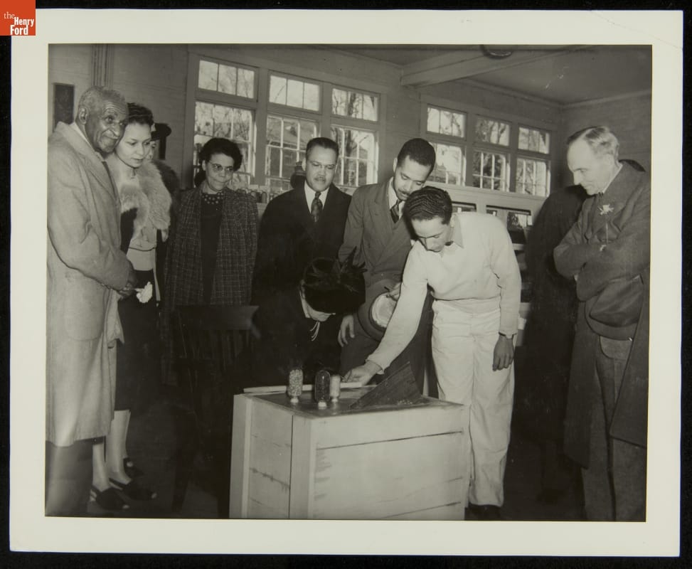 Person wearing hat sits, head tipped down toward a wooden crate, while other people look on