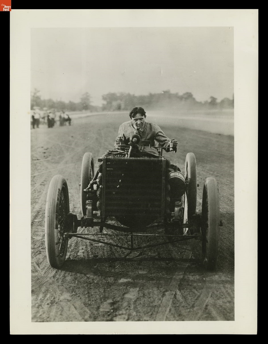 Man crouches at wheel of an open early race car on a dirt track