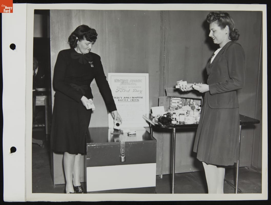 Two women with small items in their hands stand by a table and box with additional items and a sign
