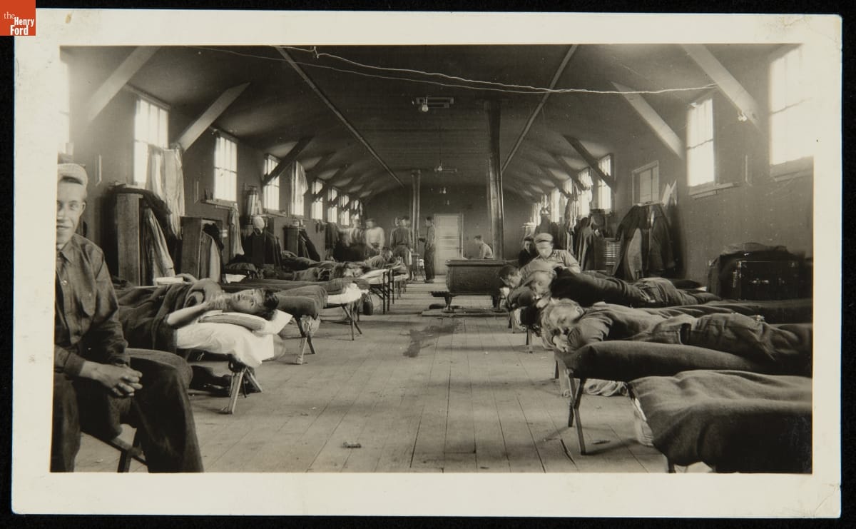 Interior of large wooden room with high windows filled with cots, some with men standing by, sitting on, or lying on them 