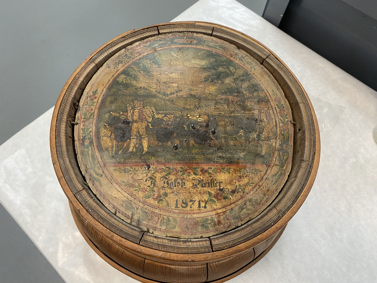 A painting of a mountain landscape with two men herding cattle is attached to the bottom of the decorative milk pail from the Gwinn Dairy Collection.