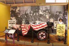 Model T covered in red and white striped bunting, in front of a photograph of women's suffrage advocates