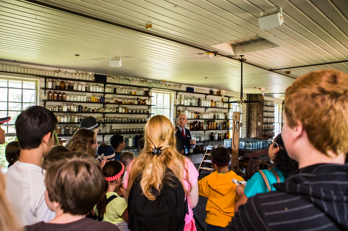 A group of children watch a man talking in a room filled with bottles and jars on shelves on the wall