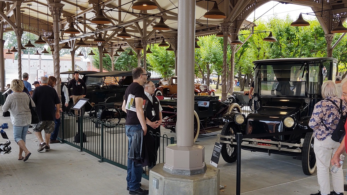Spectators look at a row of cars displayed behind barriers in an open, shed-like building