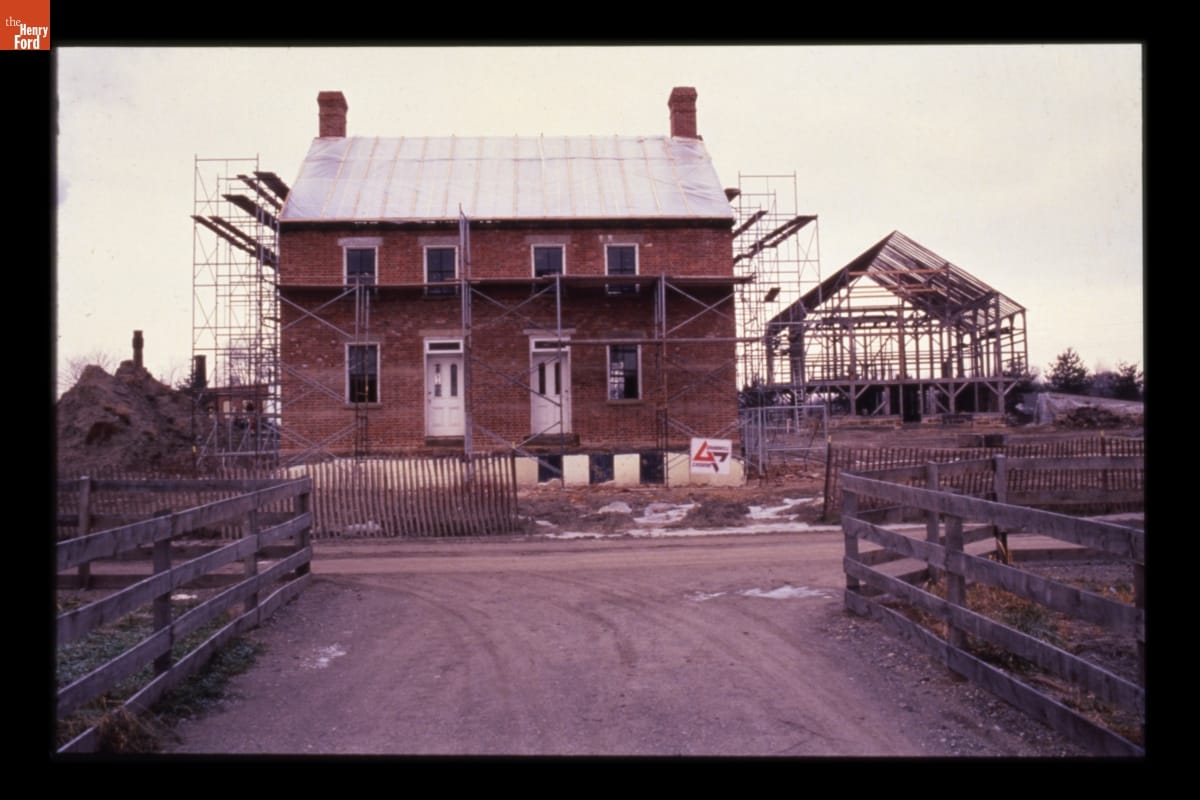 A two-story red brick house under construction, surrounded by scaffolding, with another timber-framed structure being constructed behind it