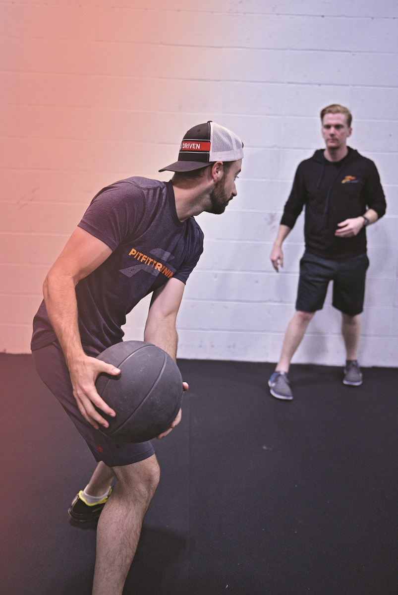 Man in shorts and t-shirt prepares to throw a medicine ball to another man in shorts and a hoodie