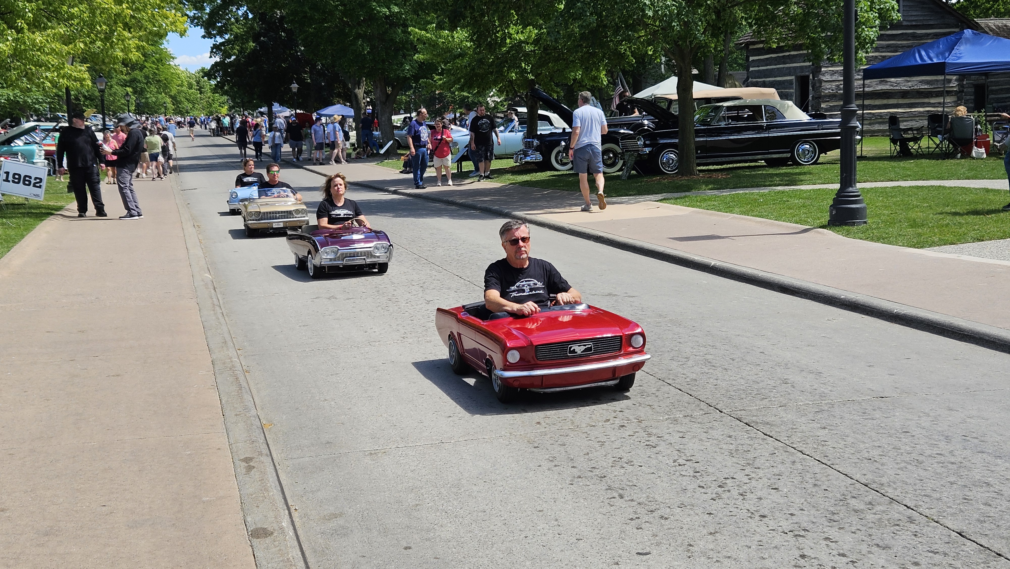 20260620_151150 A parade of Thunderbird Jr. kiddie cars led by a Mustang Jr. travels along Maple Lane during Motor Muster 2026, with young drivers enjoying the battery-powered vehicles.
