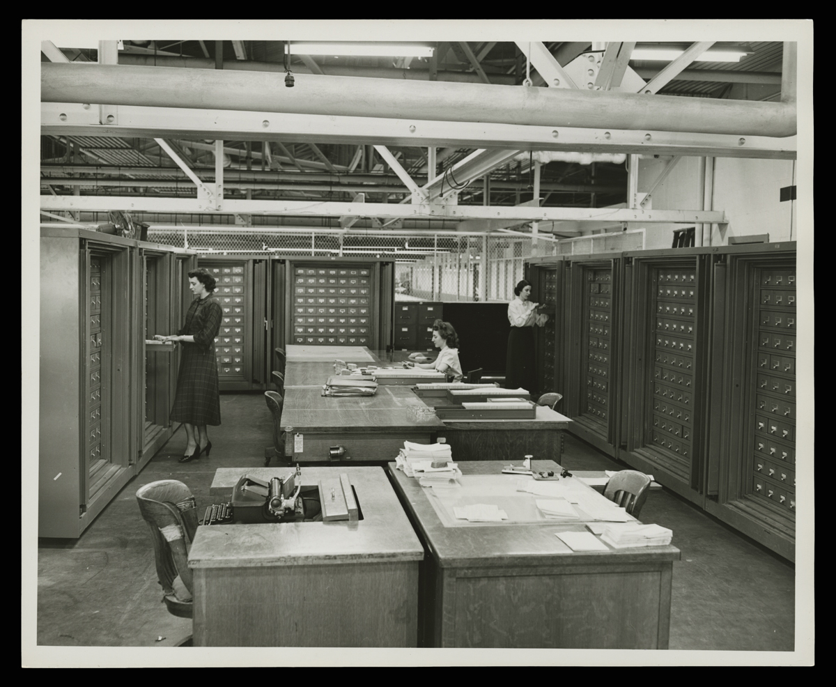 Three women in room with small filing cabinets along the walls and desks in the center