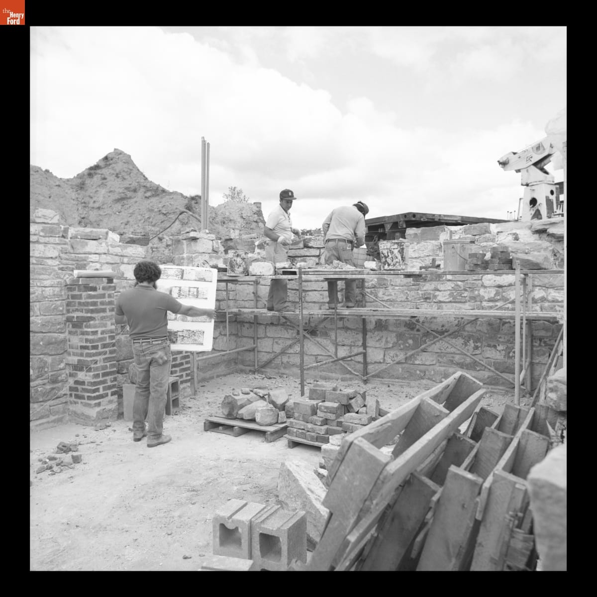Two men construct a stone block wall on a construction site, while a third man looks at a large display board he is holding that contains photographs