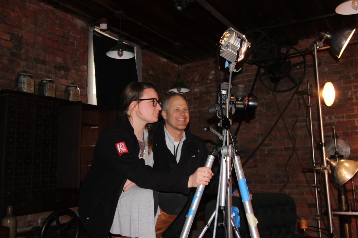 Two people behind a camera on a tripod in a brick-walled room