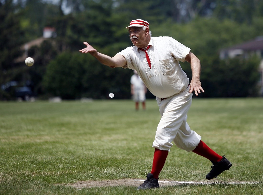 An historic base ball player in a white and red uniform throws a ball on a grassy field 