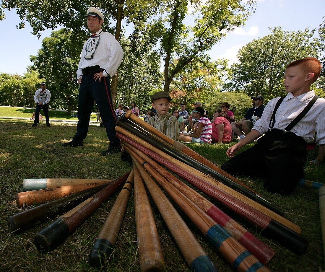 A variety of wooden bats is piled intricately in the foreground, with bat boys and historic base ball players in costume nearby, and a crowd watching the game sits behind them