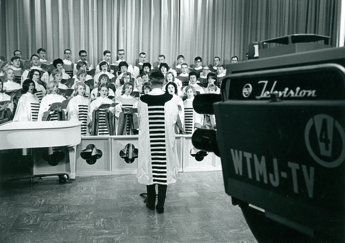 Black-and-white photo with TV camera in foreground, pointing at group of people in robes being conducted by another person in a robe