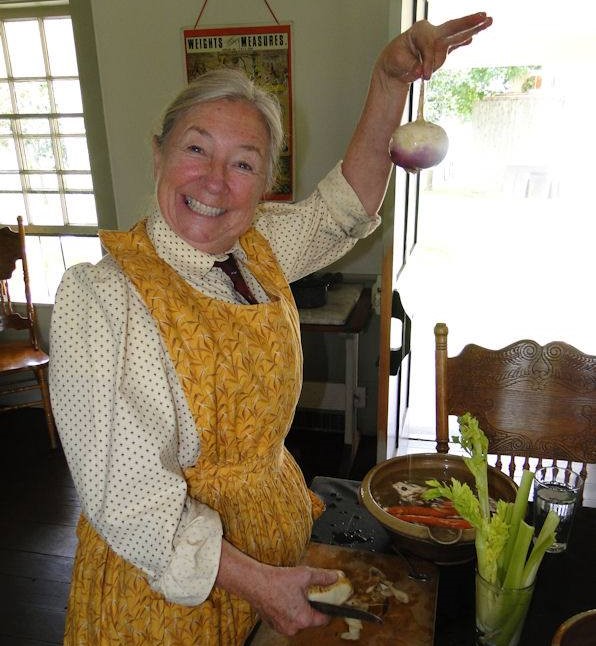 Cindy Melotti preparing food at Edison Homestead in Greenfield Village A smiling woman wearing an apron works in a kitchen, with a knife on a chopping board in one hand, holding up a turnip with the other hand
