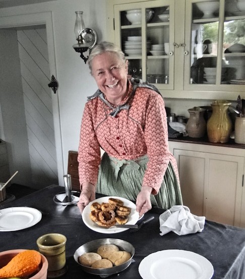 Cindy Melotti in the Ford Home kitchen in Greenfield Village A woman in a gingham dress and apron smiles and holds a plate of food in a kitchen