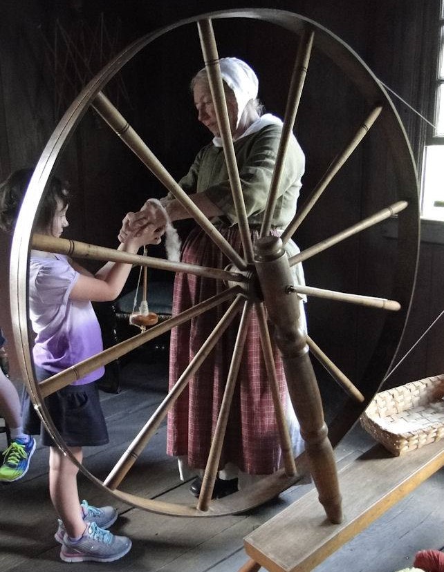 Cindy Melotti with young guest, spinning wool at Daggett Farm in Greenfield Village Woman in dress and bonnet hands a spindle to a young girl behind a large spinning wheel