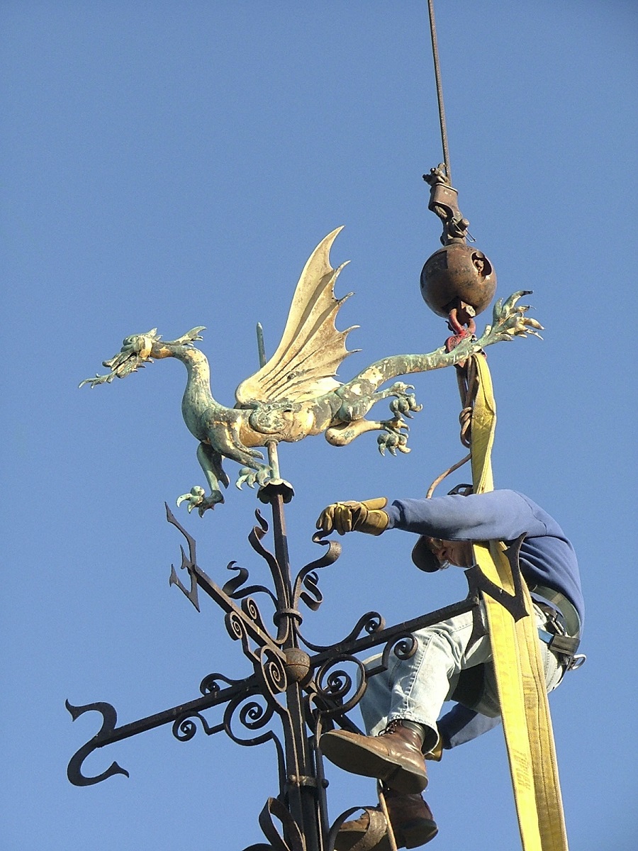 Man suspended from crane holds onto an elaborate metal weathervane in the shape of a dragon with decorative elements underneath