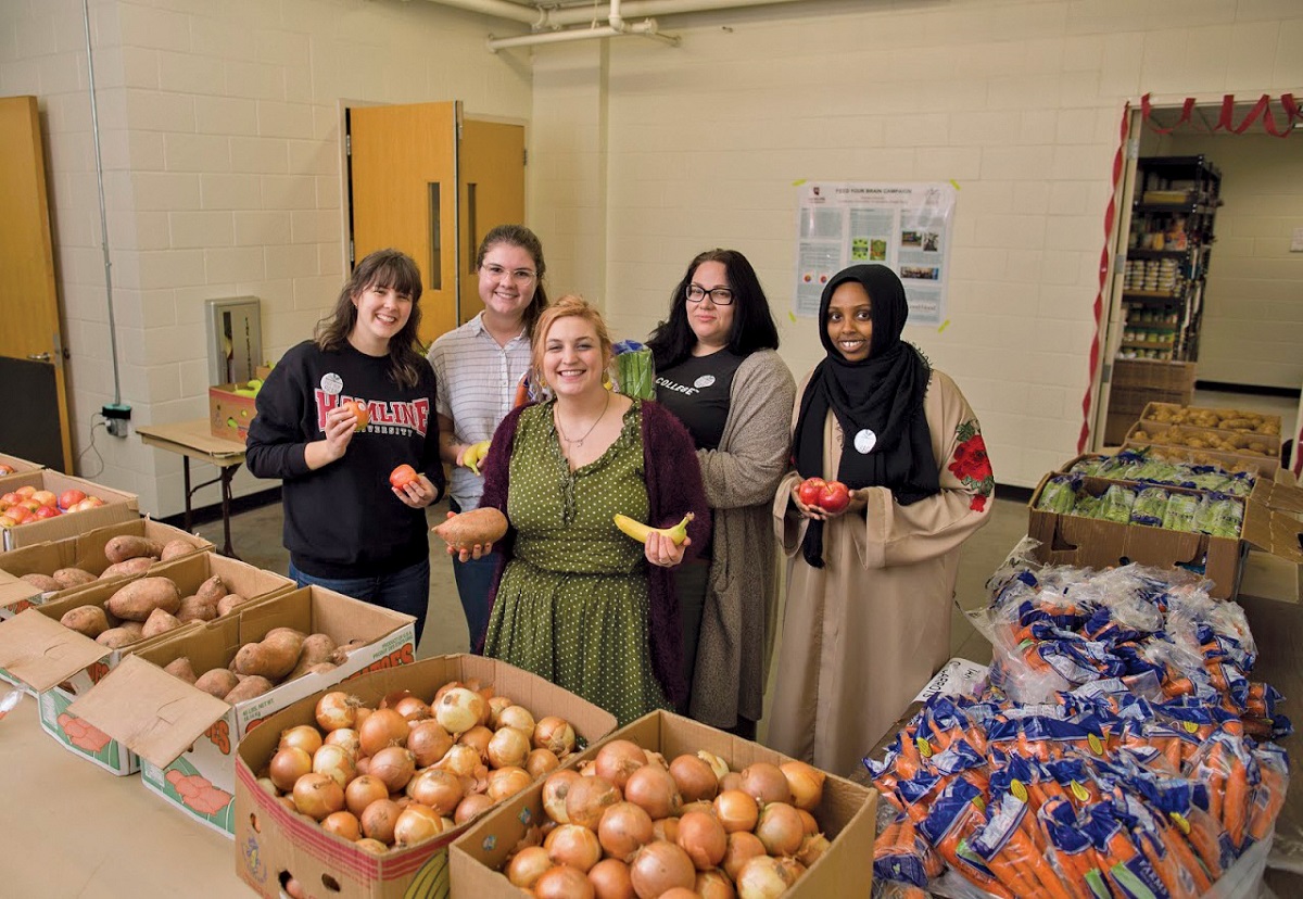 Hamline University students Five women hold produce behind tables covered in boxes full of vegetables