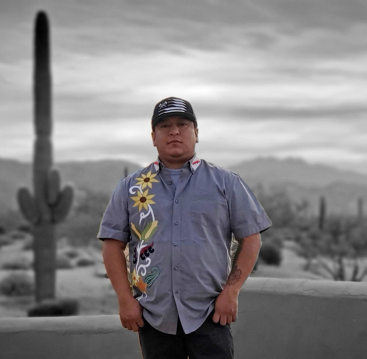 Man in short-sleeved blue button-down shirt with embroidery of flowers and vegetables along one side stands in a desert landscape with cacti and mountains