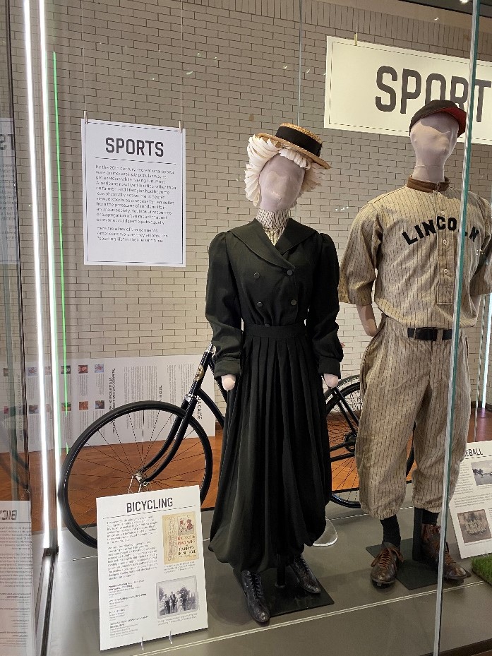 Paper wig-bedecked cyclist mannequin in the What We Wore case Mannequin wearing dress and paper wig topped with straw boater hat in display case with other items and text panels