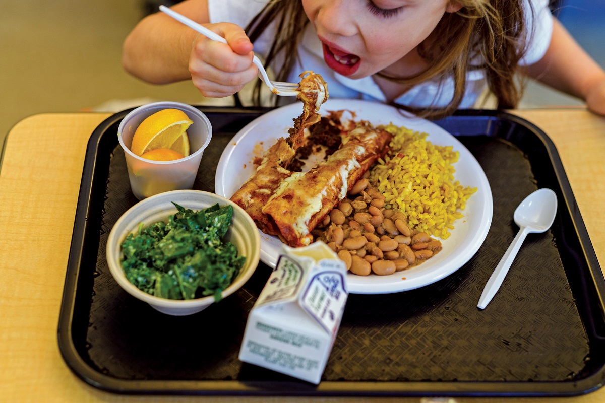 A child lifts a fork of food to her mouth behind a cafeteria tray holding enchiladas, beans, rice, orange sections, a salad, and a milk carton
