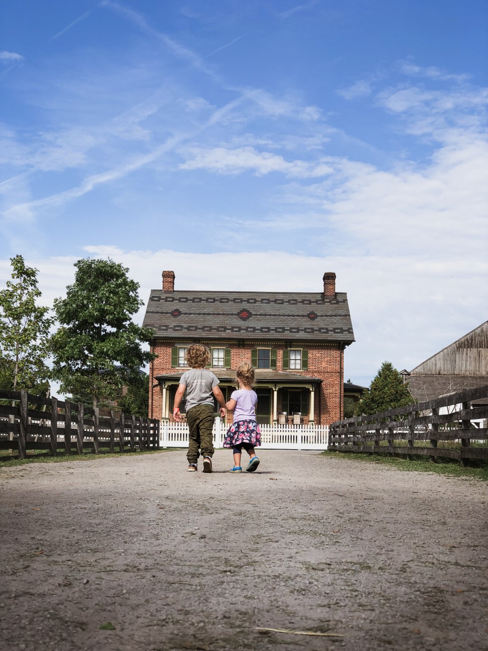 Two children walk down gravel lane between wooden fences toward a red brick farmhouse