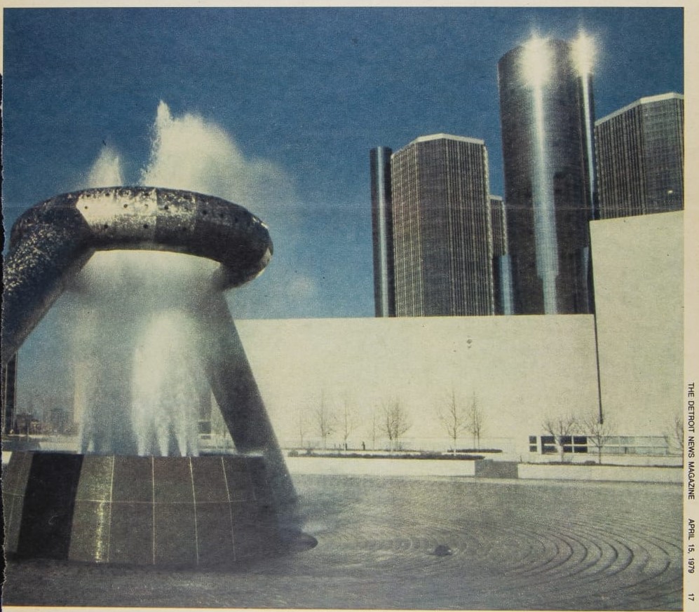 Silver fountain with large elevated silver circle through which water spouts; buildings in background