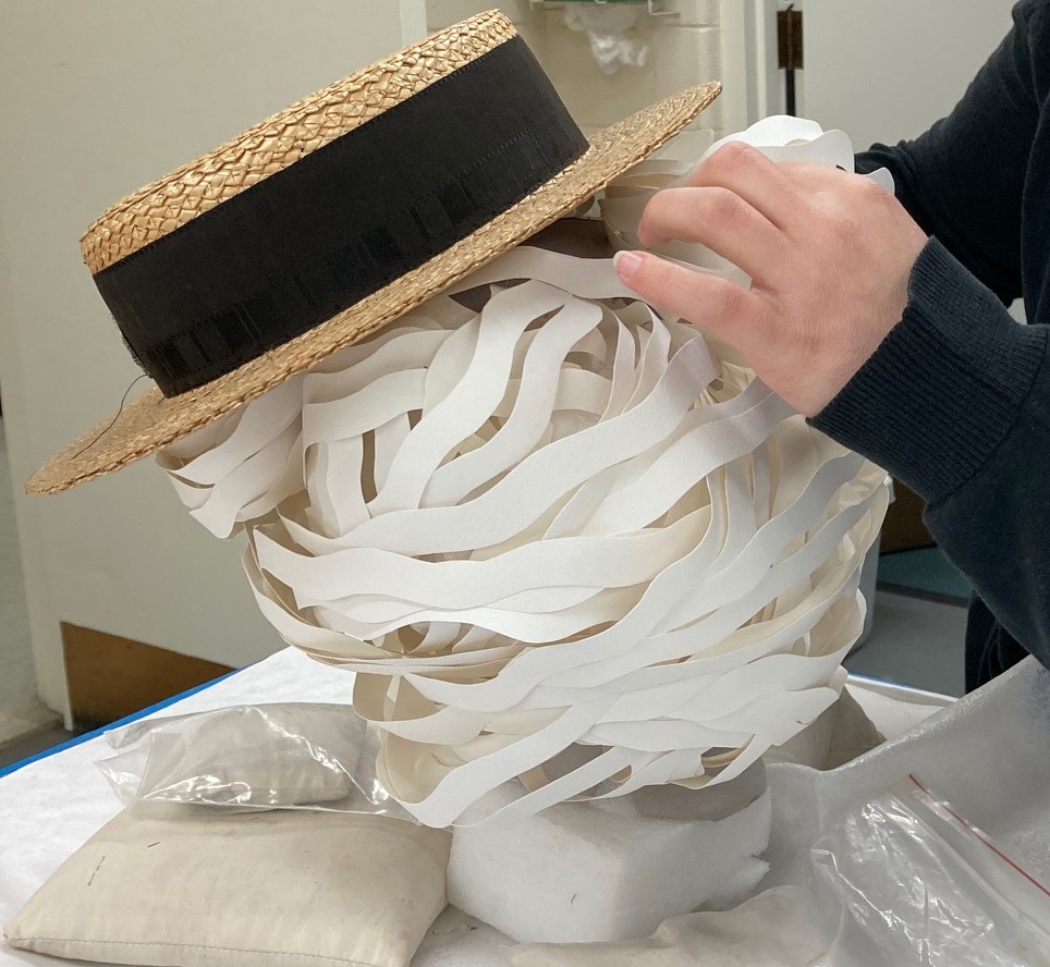 The final assembly of the wig and hat on the mannequin head White cut-paper shape with straw boater hat on top is shaped by two hands