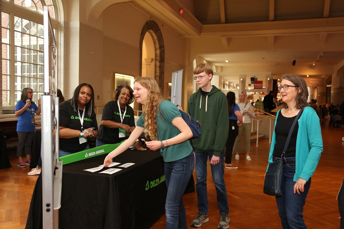 Three people look at a sign next to a table draped with a black cloth, behind which are two people in black t-shirts, in a room with other people and displays