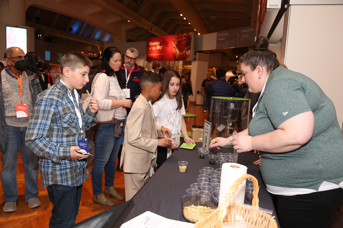 A group of children and adults stand by a table filled with small cups, behind which is a woman in a gray shirt