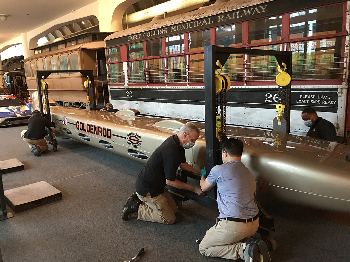 Men wearing face masks kneel around a long, low, torpedo-shaped golden car