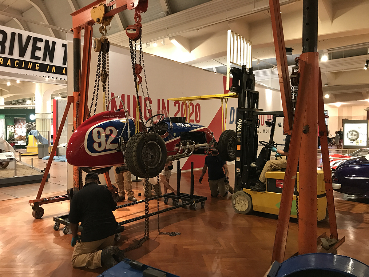 Red and blue race car suspended from a gantry and forklift with men kneeling by it