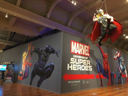 Gray wall with images and text in larger building; hanging figure of superhero in right foreground