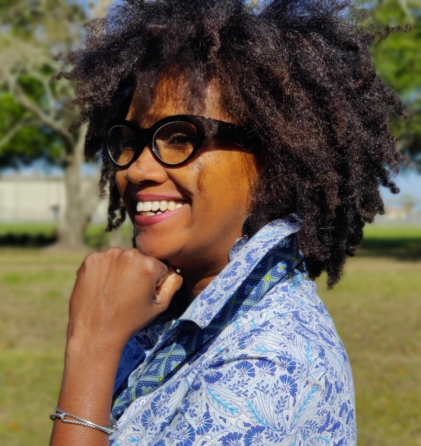 Bianca Springer, Pearland, Texas Dark-skinned woman with natural hair, glasses, and wearing a blue print shirt holds a fist to her chin and smiles