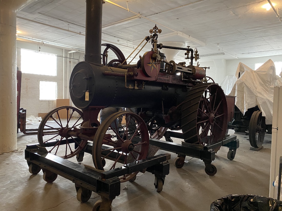 A steam traction engine is lifted onto custom-built dollies A large piece of wheeled, black machinery sits on multiple dollies in a bright room