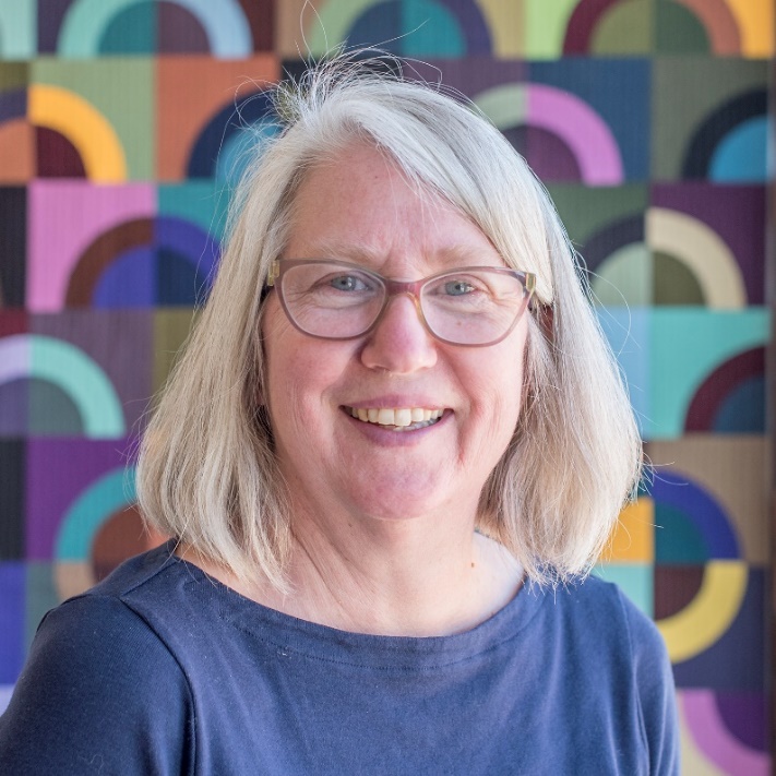 Lynn Carson Harris, Chelsea, Michigan Light-skinned woman with blonde hair and glasses, wearing blue top, in front of a colorful backdrop