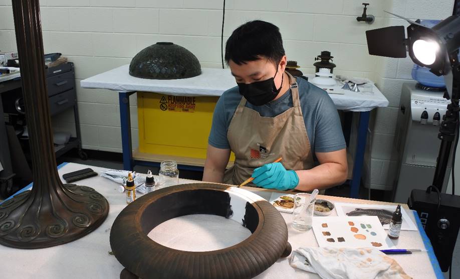 Conservator Cuong Nguyen painting the 3D-printed lamp base Man wearing face mask sits at a table covered with bottles, jars, and papers, painting a round lamp base brown