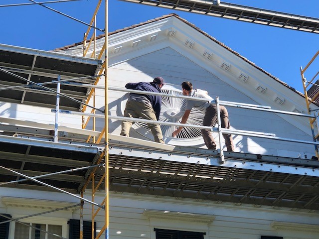 Two men on scaffolding hold a large wooden architectural element on a building façade