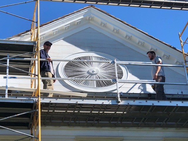 Two men on scaffolding stand on either side of a large wooden architectural element on a building façade