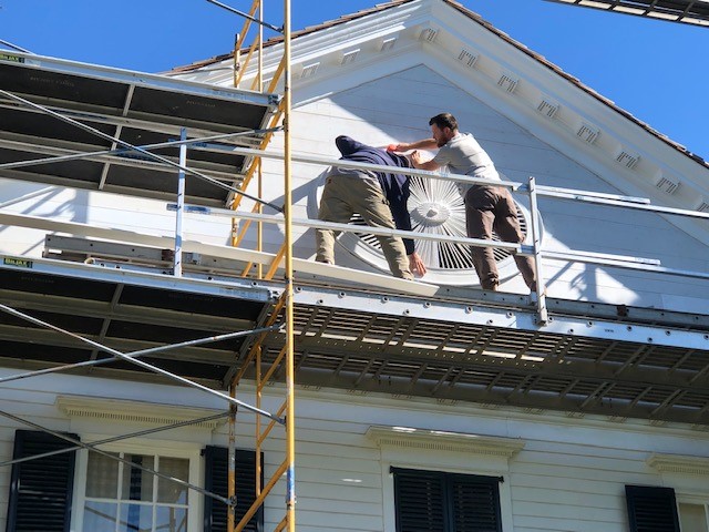 Two men on scaffolding hold a large wooden architectural element on a building façade