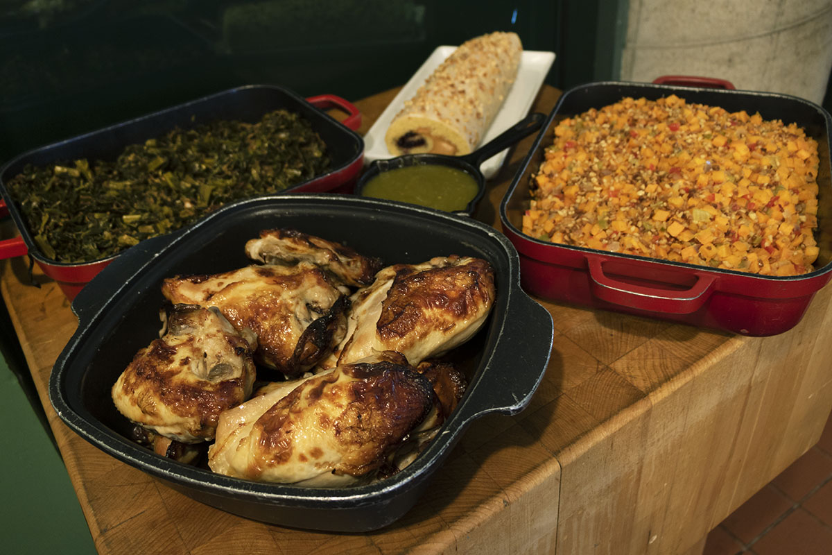 Dishes containing chicken, greens, mixed vegetables, a green sauce, and a rolled cake