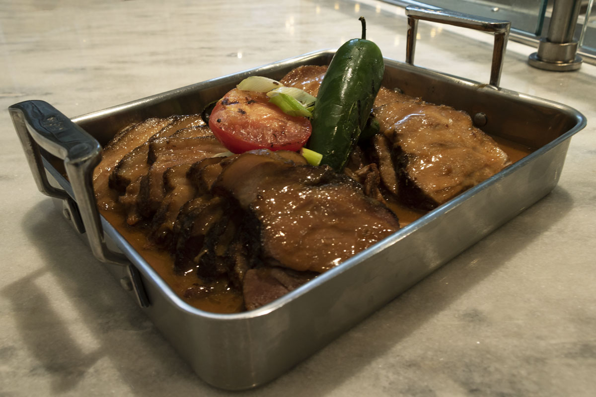 Silver platter filled with meat in a brown sauce, topped with tomato slices and jalapeno peppers; text label behind dish