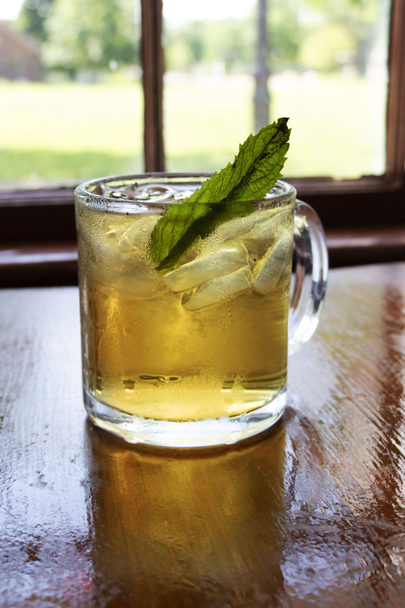 Eagle Tavern's Mint Iced Tea Clear glass mug filled with ice and amber-colored liquid, garnished with a mint leaf, sitting on a wooden table in front of a window
