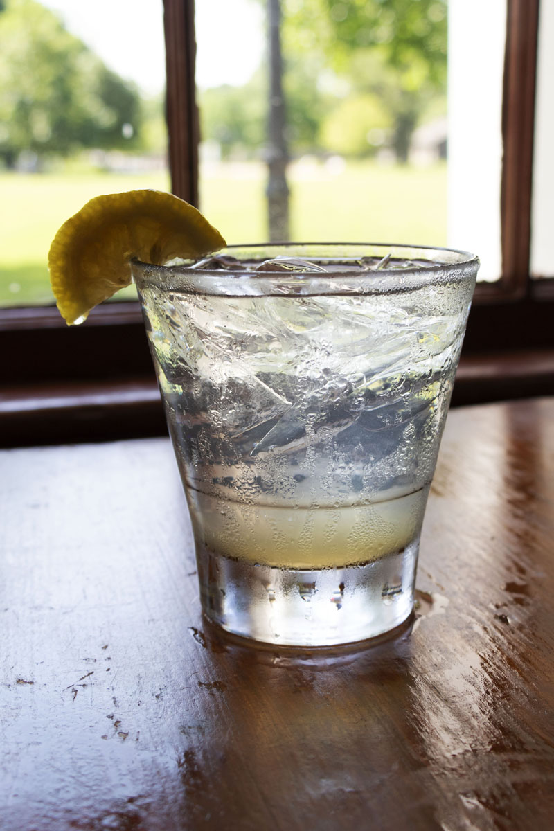 Lemon & Ginger Shrub Clear glass with lemon on rim, filled with ice and beverage, sitting on a wooden table with a window in the background