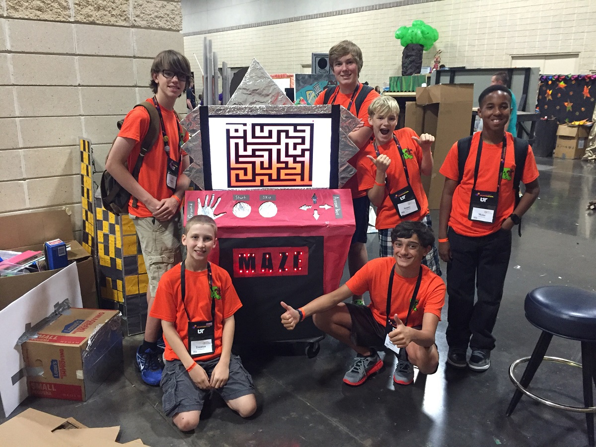 Group of children stand and kneel around an arcade game made out of cardboard