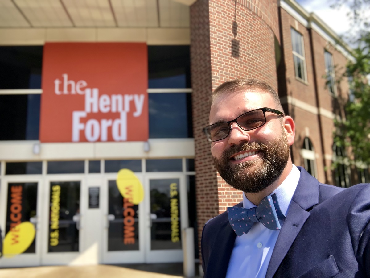 Man in blue suit and bow tie takes a selfie in front of a building with large logo reading "The Henry Ford"