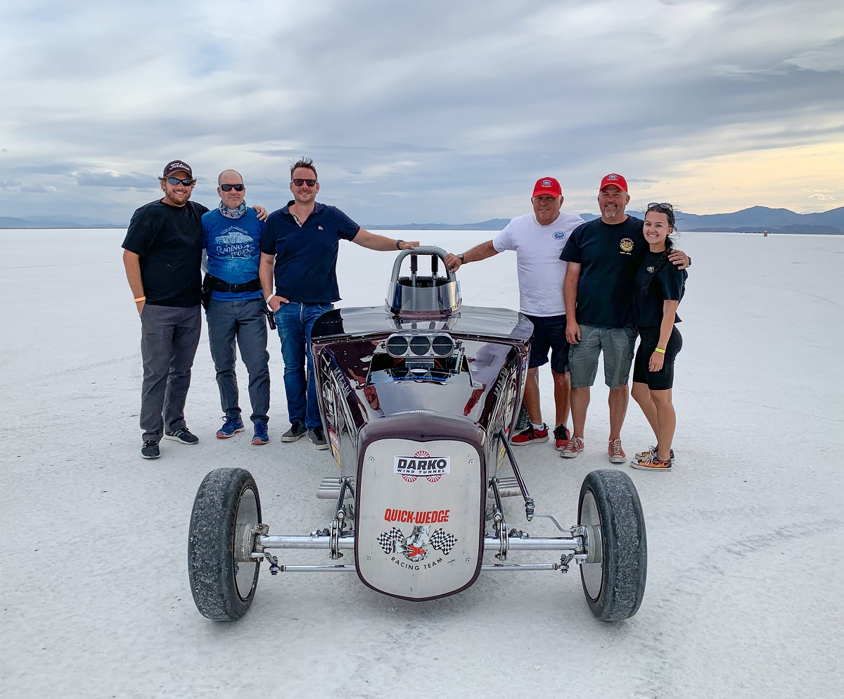 Six people stand around a race car on a salt plain with mountains in the background