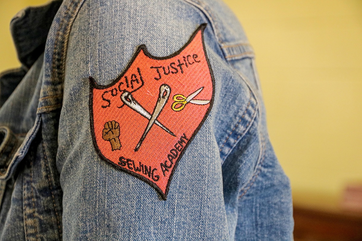 SJSA patch Red, shield-shaped patch with text and images of two crossed sewing needles, an upraised brown fist, and a pair of scissors, on the sleeve of a jean jacket