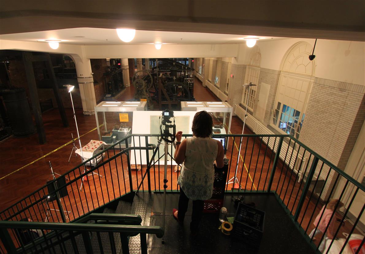 Person with camera on landing at top of staircase, looking down over railing toward a large tilted board with a textile on it